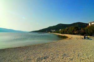 Nyugodt strand aranyszínű homokkal és nyugodt, tiszta vízzel a ragyogó kék ég alatt. Néhány ember heverészik néhány napernyő alatt a parton. A távolban zöld dombok és néhány szétszórt ház látható.