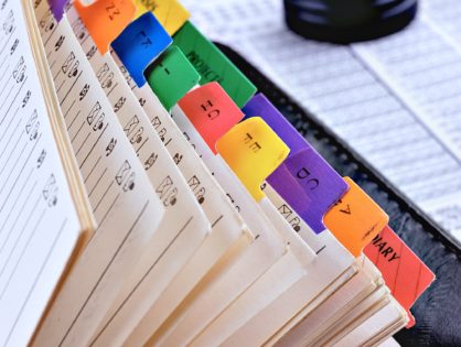 Közeli kép egy megnyitott fájlmappáról, színes indexekkel ábécé sorrendben. A háttérben egy fából készült bélyegző látható. Az anyakönyvek egyedi betűkkel vannak felcímkézve, és papíralapú dokumentumokat tartalmaznak, a regiszterek színes széleit kiemelve.