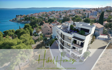 Ein modernes weißes Apartmenthaus mit Blick auf die Adria, umgeben von Grün und Häusern in einer kroatischen Küstenstadt unter einem klaren blauen Himmel. Der überlagerte Text lautet "Hike Homes - Traum- & Ferienimmobilien in Kroatien. - Luxus Meerblick 77m² Ensemble mit 2 Schlafzimmern, Terrasse und Südausrichtung