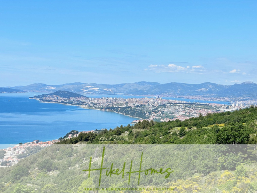 Exklusive 380m² Meerblick & Luxus in absoluter Alleinlage mit Privatspähre + Pool, Sommerküche, … - Ein Panoramablick auf eine Küstenstadt mit blauem Meer, grünen Hügeln und entfernten Bergen unter einem klaren blauen Himmel. Die Stadt erstreckt sich entlang der Küstenlinie, mit Gebäuden und Grünflächen sichtbar. (44524)