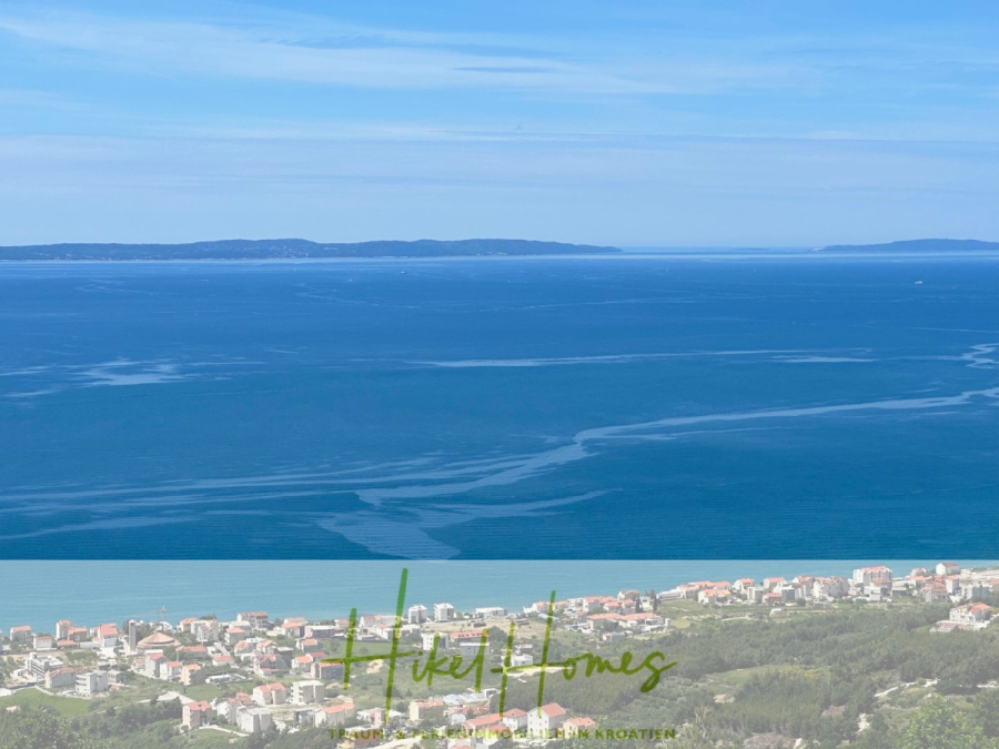 Exklusive 380m² Meerblick & Luxus in absoluter Alleinlage mit Privatspähre + Pool, Sommerküche, … - Blick auf eine Küstenstadt mit rotgedeckten Gebäuden, die im Vordergrund von Grün umgeben sind, mit Blick auf ein weites blaues Meer und entfernte Inseln unter einem klaren blauen Himmel. Der Schriftzug "Niki Homes" erscheint am unteren Rand. (44526)