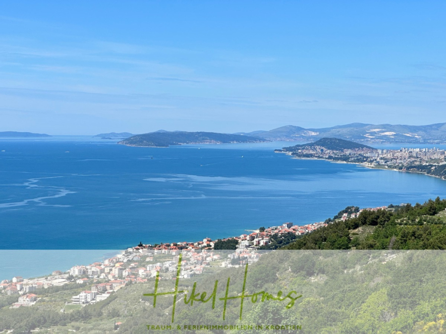 Exklusive 380m² Meerblick & Luxus in absoluter Alleinlage mit Privatspähre + Pool, Sommerküche, … - Eine Küstenstadt mit weißen Häusern erstreckt sich entlang der Küste, umgeben von üppig grünen Hügeln und blauem Meer, mit entfernten Bergen unter einem klaren Himmel. Der Schriftzug "Hikel Homes" ist am unteren Rand eingeblendet. (44525)