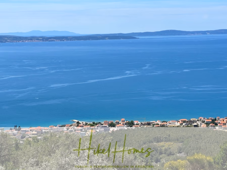 Exklusive 380m² Meerblick & Luxus in absoluter Alleinlage mit Privatspähre + Pool, Sommerküche, … - Panoramablick auf eine Küstenstadt mit einer Ansammlung von Gebäuden in Ufernähe. Das weite blaue Meer erstreckt sich unter klarem Himmel bis zum Horizont, und über das Wasser hinweg sind ferne Landmassen zu erkennen. Im Vordergrund steht die Vegetation. (41906)