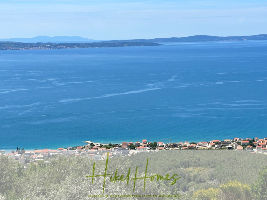 Exklusive 380m² Meerblick & Luxus in absoluter Alleinlage mit Privatspähre + Pool, Sommerküche, … - Ein Küstendorf mit roten Dächern liegt an einem ruhigen blauen Meer mit fernen Hügeln auf der anderen Seite des Wassers unter einem klaren Himmel. Im Vordergrund sind Grünflächen und Bäume zu sehen. Der Text "Heled Homes" erscheint am unteren Rand. (44528)