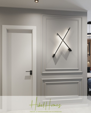 Ein moderner, minimalistischer Flur mit einer weißen Tür, dekorativen Wandleisten und einer schwarzen, X-förmigen Wandleuchte. Die Farbpalette ist neutral mit Schattierungen von Grau und Weiß. Der Schriftzug "Hiked Homes" ist am unteren Rand sichtbar. - Meerblick 76m² Neubauwohnung mit 2 Schlafzimmer, Balkon und Südausrichtung