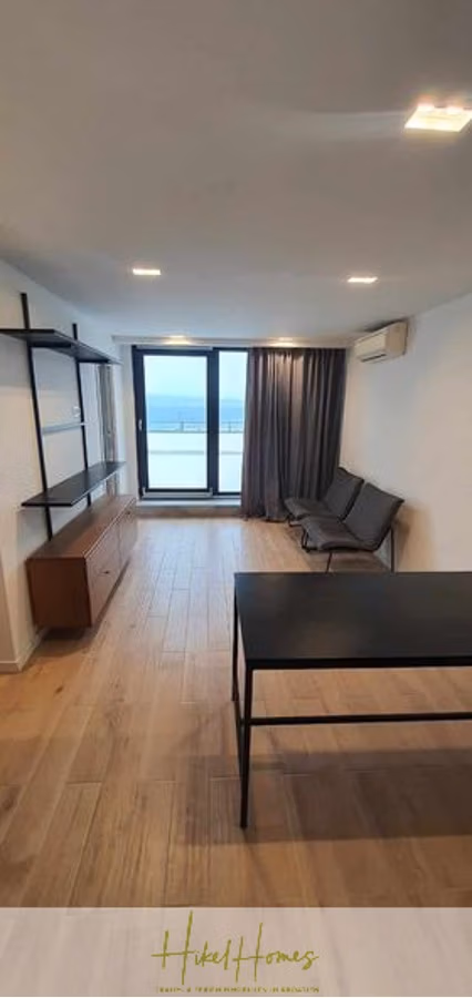 Charmantes 1-Zimmer-Appartement mit Meerblick im Herzen von Split mit Dachterrasse - Minimalistisches Wohnzimmer mit Parkettboden, schwarzem Tisch und dunklen Stühlen. Links befinden sich ein Wandregal und ein Sideboard. Große Fenster mit grauen Vorhängen bieten Ausblick, darüber befindet sich eine Klimaanlage. (42223)