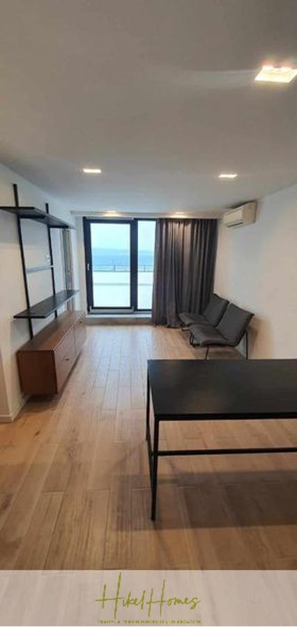Charmantes 1-Zimmer-Appartement mit Meerblick im Herzen von Split mit Dachterrasse - Modernes Wohnzimmer mit Holzboden, schwarzem Schreibtisch, minimalistischem schwarzem Sofa, Regalen und großen Glastüren, die auf einen Balkon mit Meerblick führen. An der rechten Wand befinden sich graue Vorhänge und eine Klimaanlage. (45409)