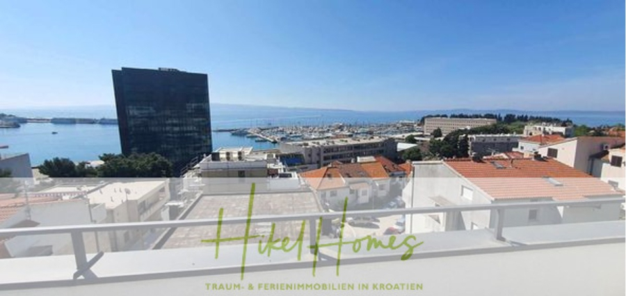 Charmantes 1-Zimmer-Appartement mit Meerblick im Herzen von Split mit Dachterrasse - Blick von einem Balkon auf Dächer, einen Hafen mit Booten, ein hohes Gebäude aus dunklem Glas und das Meer im Hintergrund unter einem klaren blauen Himmel. Der Text "Hikel Homes" ist am unteren Rand eingeblendet. (45406)