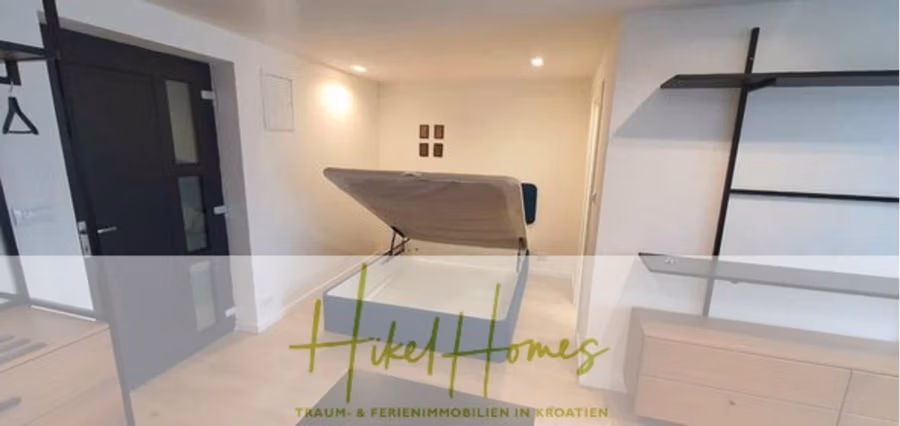 Charmantes 1-Zimmer-Appartement mit Meerblick im Herzen von Split mit Dachterrasse - Ein kompakter Raum mit einem hochgeklappten Schrankbett an einer hellen Wand. Der Raum ist mit Holzboden und minimalistischer Möblierung ausgestattet, darunter eine Kommode und ein offenes Regal. Die Tür ist dunkel gehalten. Das Bild zeigt ein transparentes Banner mit Text. (42222)