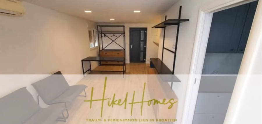 Charmantes 1-Zimmer-Appartement mit Meerblick im Herzen von Split mit Dachterrasse - Modernes Apartment-Interieur mit minimalistischem Design, weißen Wänden, hellem Holzboden und grauen Möbeln. Ein an der Wand montierter Fernseher, ein Regal und ein kleiner schwarzer Tisch sind sichtbar. Ein Schild mit der Aufschrift „Hikel Homes, Traum- & Ferienimmobilien in Kroatien“ steht auf dem Foto. (42226)