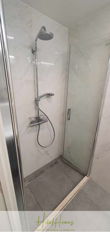 Charmantes 1-Zimmer-Appartement mit Meerblick im Herzen von Split mit Dachterrasse - Eine moderne Dusche mit einer an der Wand montierten verstellbaren Kopfbrause, einer Handbrause und einer Glastür. Die Wände sind weiß und der Boden ist grau gefliest. Das Logo von Hitt Homes ist am Boden sichtbar. (45413)