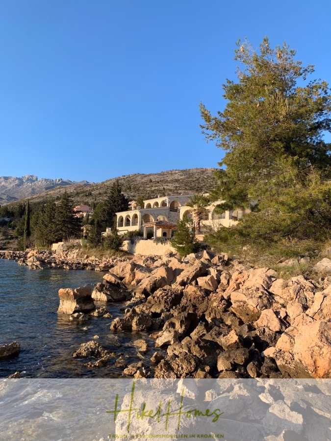 1160m² in erste Reihe zum Meer / Ideal zur Bebauung mit 2 Villen in erster und zweiter Reihe - Eine sonnendurchflutete Küstenvilla mit Bogenfenstern liegt eingebettet zwischen Bäumen und felsigen Ufern und bietet Blick auf das klare, blaue Meer. Im Hintergrund sind schroffe Hügel unter einem strahlend blauen Himmel zu sehen. (42372)