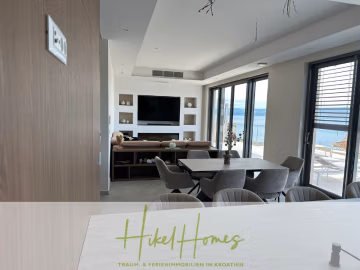 Moderner Wohn- und Essbereich mit neutralen Farbtönen, großen Fenstern mit Blick auf das Meer und modernen Möbeln. Der Raum ist mit einem eingebauten TV-Gerät und Wandregalen ausgestattet. Der Schriftzug "Hikel Homes" ist am Boden sichtbar. - Luxus pur! Jetzt einziehen und Sommer genießen! Schlüsselfertige Meerblick Villa, 3SZ, Pool, …