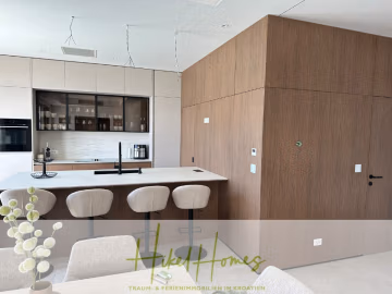 Moderne Küche mit hellen Holzschränken, einer weißen Insel mit vier beigen Barhockern, einem eingebauten Ofen, Regalen mit Glaswaren und einem minimalistischen Dekor. Der Text "Hikel Homes" ist am unteren Rand des Bildes eingeblendet. - Luxus pur! Jetzt einziehen und Sommer genießen! Schlüsselfertige Meerblick Villa, 3SZ, Pool, …