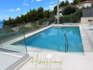 Ein modernes Freibad mit Glasumrandung, umgeben von Grün und mit Blick auf das Meer. Im Hintergrund sind einige Häuser und Pinienbäume unter blauem Himmel zu sehen. Das Logo von "Hikel Homes" erscheint am unteren Rand. - Luxus pur! Jetzt einziehen und Sommer genießen! Schlüsselfertige Meerblick Villa, 3SZ, Pool, …