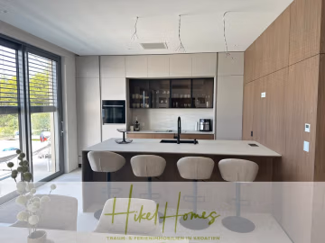 Moderne Küche mit beigen Schränken, einer großen Insel mit vier grauen Drehstühlen, schwarzen Armaturen, eingebautem Backofen und einer Kaffeemaschine. Große Fenster sorgen für natürliches Licht. "Hike Homes"-Logo am Boden. - Luxus pur! Jetzt einziehen und Sommer genießen! Schlüsselfertige Meerblick Villa, 3SZ, Pool, …