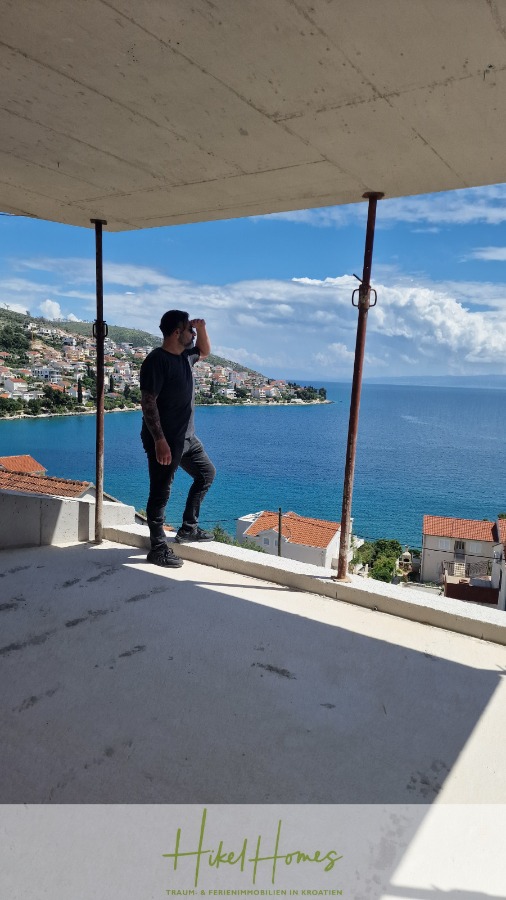 Luxus Meerblick 77m² Ensemble mit 2 Schlafzimmern, Terrasse und Südausrichtung – Einziehen 2025! - Ein schwarz gekleideter Mann steht auf dem Balkon eines unvollendeten Gebäudes und blickt auf eine Küstenstadt und das blaue Meer unter einem teilweise bewölkten Himmel. Im Hintergrund sind Dächer und Hügel zu sehen. (46467)