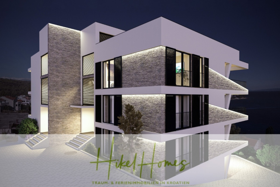 Luxus Meerblick 77m² Ensemble mit 2 Schlafzimmern, Terrasse und Südausrichtung – Einziehen 2025! - Ein modernes, mehrstöckiges Gebäude mit großen Fenstern und leuchtenden Außenlichtern bei Nacht, mit Blick auf das Wasser. Der Text "Hikel Homes Traum- & Ferienimmobilien in Kroatien" ist am unteren Rand eingeblendet. (46457)