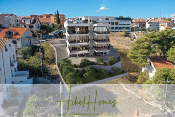 Ein modernes, mehrstöckiges Apartmentgebäude mit großen Balkonen steht auf einem Hanggrundstück inmitten von Häusern mit roten Ziegeldächern in einer kroatischen Küstenstadt. Das Bild enthält den Text: „Hikel Homes – Traum- & Ferienimmobilien in Kroatien.“. - Luxus Meerblick 77m² Ensemble mit 2 Schlafzimmern, Terrasse und Südausrichtung – Einziehen 2025!