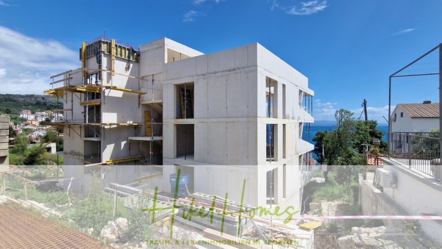 Luxus Meerblick 77m² Ensemble mit 2 Schlafzimmern, Terrasse und Südausrichtung – Einziehen 2025! - Ein modernes, mehrstöckiges Gebäude im Bau mit Sichtbetonwänden, Gerüsten und einem blauen Himmel über sich. Im Hintergrund sind das Meer und andere Häuser zu sehen. (46459)