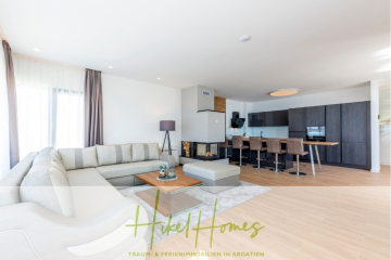 Modernes offenes Wohnzimmer und Küche mit einem großen hellgrauen Sektionssofa, einem Couchtisch aus Holz, einem langen Esstisch mit braunen Stühlen, dunklen Schränken und hellem Holzfußboden. Der helle, geräumige Raum wird von Sonnenlicht durchflutet. - Sofort Einziehen oder attraktive Rendite! Meerblick, 4 SZ, Pool, Solar, Garage, …