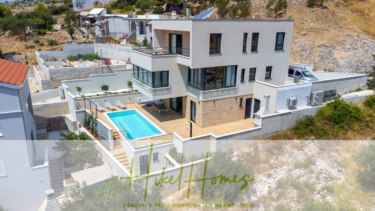 Eine moderne weiße Villa mit großen Fenstern, Balkonen und einem privaten Swimmingpool, die auf einem Hügel mit angelegten Terrassen und umliegenden Grünflächen liegt. Der Text "Hike Homes" ist am unteren Rand des Bildes zu sehen. - Sofort Einziehen oder attraktive Rendite! Meerblick, 4 SZ, Pool, Solar, Garage, …