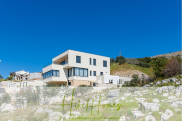Ein modernes, zweistöckiges weißes Haus mit großen Fenstern steht auf einem felsigen Hügel unter einem klaren blauen Himmel. Das Grundstück ist von Grün umgeben, und der Text "Hikel Homes" ist am unteren Rand des Bildes eingeblendet. - Sofort Einziehen oder attraktive Rendite! Meerblick, 4 SZ, Pool, Solar, Garage, …