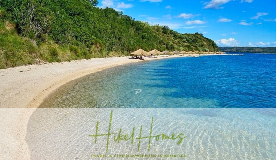 Neubau Meerblick 270m vom Meer entfernt – Hochwertigen Ausstattung – Fertigstellung Ende 2025 - Strandszene mit klarem, blauem Wasser und einem Sandstrand mit Sonnenschirmen und Liegestühlen. Dichtes Grün säumt den Strand, und im Hintergrund erhebt sich unter einem überwiegend klaren, blauen Himmel ein grasbewachsener Hügel. (42605)