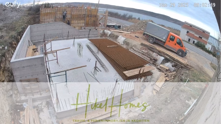 Neubau Meerblick 270m vom Meer entfernt – Hochwertigen Ausstattung – Fertigstellung Ende 2025 - Baustelle mit Betonfundament und Bewehrungsstäben. Arbeiter errichten ein Holzgerüst. Ein orangefarbener LKW parkt in der Nähe. Im Hintergrund ist ein nahegelegenes Gewässer zu sehen. Textüberlagerung: „Hikel Homes – Traum- & Ferienimmobilien in Kroatien.“. (42612)