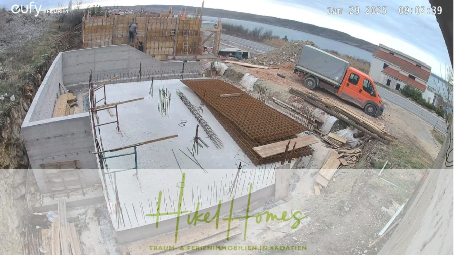 Neubau Meerblick 270m vom Meer entfernt – Hochwertigen Ausstattung – Fertigstellung Ende 2025 - Eine Baustelle mit Betonwänden und Stahlverstärkungen, Arbeitern auf Gerüsten, einem orangefarbenen Lastwagen, der in der Nähe geparkt ist, und einem See im Hintergrund. Der Text am unteren Rand lautet "Hikel Homes" und "Immobilien in Kroatien". (46245)