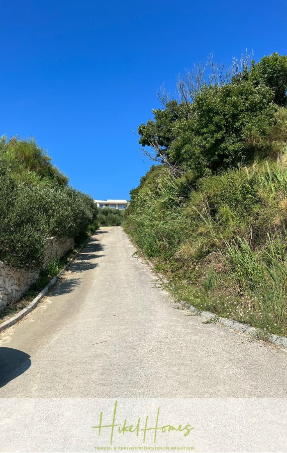 Neubau Meerblick 270m vom Meer entfernt – Hochwertigen Ausstattung – Fertigstellung Ende 2025 - Eine schmale, gepflasterte Straße führt zwischen grünen, buschbewachsenen Ufern unter einem klaren, blauen Himmel sanft bergauf. Auf der Spitze des Hügels ist ein kleines Gebäude mit einem roten Dach zu sehen. (46237)