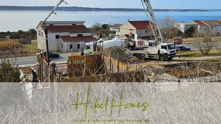 Neubau Meerblick 270m vom Meer entfernt – Hochwertigen Ausstattung – Fertigstellung Ende 2025 - Baustelle in der Nähe eines Gewässers, mit Arbeitern, Lastwagen und Maschinen, die Beton gießen. Im Hintergrund sind Wohngebäude und eine Straße zu sehen. Das Logo von "Hudil Homes" erscheint am unteren Rand des Bildes. (46244)