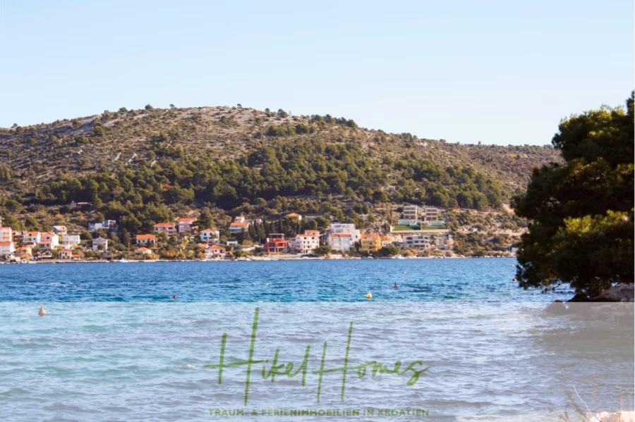 Luxus Villa Sunce / gegenüber Marina Frapa / Meerblick + direkter Meerzugang, Pool, mit 391m² WFL - Eine Küstenlandschaft mit klarem, blauem Wasser und Häusern inmitten üppiger, grüner Hügel. Mehrere farbenfrohe Häuser stehen verstreut am Ufer. Im Vordergrund sind ein Baum und ein Wasserzeichen mit der grünen Aufschrift „Hotel Homes“ zu sehen. (42022)