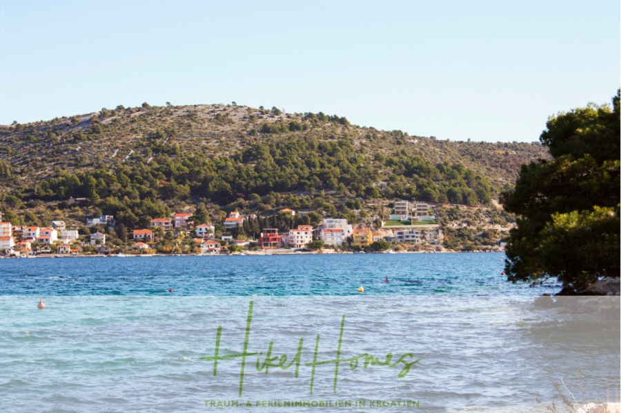 Luxus Villa Sunce / gegenüber Marina Frapa / Meerblick + direkter Meerzugang, Pool, mit 391m² WFL - Eine Küstenszene mit klarem blauen Wasser im Vordergrund, bunten Häusern entlang der Küste, grünen Hügeln im Hintergrund und dem Logo "Hikel Homes" mit Text über Immobilien in Kroatien am unteren Rand. (45119)