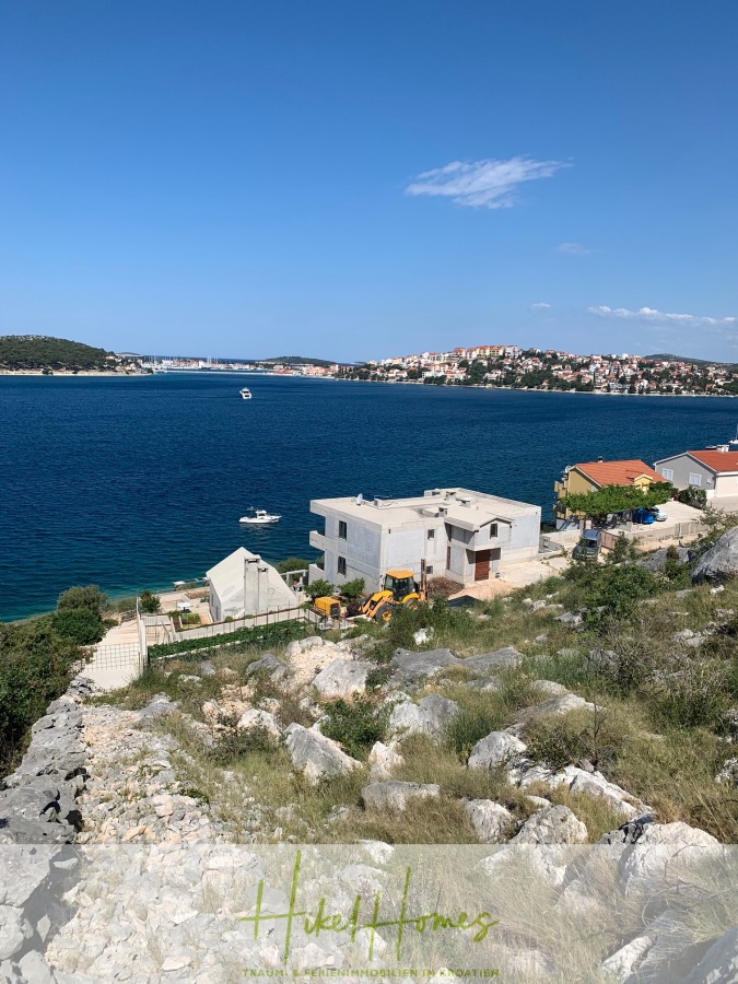Luxus Villa Sunce / gegenüber Marina Frapa / Meerblick + direkter Meerzugang, Pool, mit 391m² WFL - Blick auf eine Küstenstadt mit blauem Meer und Booten, modernen weißen Häusern in Küstennähe, felsigem Gelände mit Gras im Vordergrund und einer Ansammlung von Gebäuden auf dem entfernten Hügel unter einem klaren blauen Himmel. (45122)