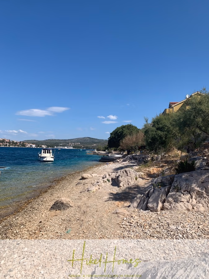 Luxus Villa Sunce / gegenüber Marina Frapa / Meerblick + direkter Meerzugang, Pool, mit 391m² WFL - Ein malerischer Blick auf einen felsigen Strand mit einem Boot auf dem ruhigen blauen Meer unter klarem Himmel. Bäume und ein gelbes Gebäude sind auf der rechten Seite zu sehen. Im Hintergrund sind Hügel und eine Küstenstadt zu sehen. (42028)