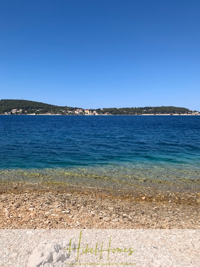 Luxus Villa Sunce / gegenüber Marina Frapa / Meerblick + direkter Meerzugang, Pool, mit 391m² WFL - Ein klarer, blauer Himmel über einer ruhigen Küstenlandschaft mit einem felsigen Strand im Vordergrund und einem ruhigen Meer, das sich bis zu einer fernen, baumbestandenen Küste erstreckt. Am unteren Bildrand ist das Logo „Heidi Homes“ zu sehen. (42027)