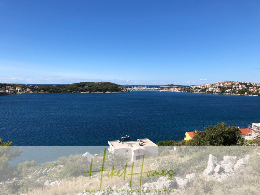 Luxus Villa Sunce / gegenüber Marina Frapa / Meerblick + direkter Meerzugang, Pool, mit 391m² WFL - Ein hellblauer See oder ein Meer mit sanften Wellen, umgeben von grünen Hügeln und einer Küstenstadt mit verstreuten Häusern unter einem klaren blauen Himmel. Im Vordergrund sind weiße Gebäude und felsiges Terrain zu sehen. (45136)