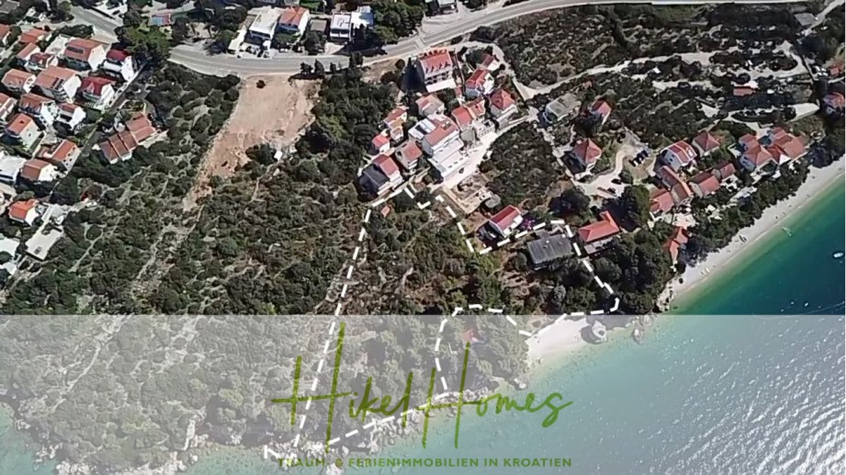 Luftaufnahme eines Küstendorfes mit Häusern entlang einer kurvenreichen Straße und einem kleinen Strand neben klarem türkisfarbenem Wasser. Das Bild enthält das Logo "HeKel Homes" mit dem Text "IHR PARTNER FÜR FERIENIMMOBILIEN IN KROATIEN". - 7448qm in erste Reihe zum Meer / Ideal zur Bebauung oder touristischen Betrieb (Hotel / Appartement)