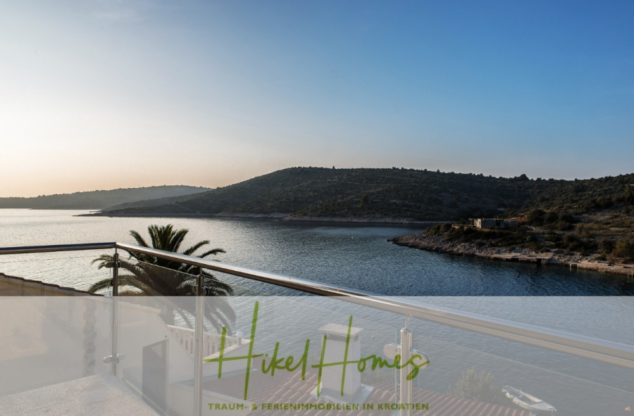 5 Sterne Luxus Villa Christina – 250m² WFL in 1. Reihe zum Meer/ 4 SZ / Neubau / Sofort bezugsfertig - Blick von einem Balkon auf eine ruhige Bucht mit klarem Wasser, einer Palme und sanften grünen Hügeln bei Sonnenuntergang. Entlang des Balkons verläuft ein Glasgeländer. Der Text am unteren Rand lautet "Hikel Homes" und "Feriendomizile in Kroatien". (46312)