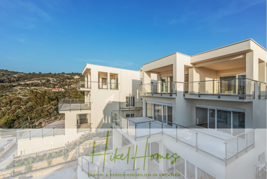 5 Sterne Luxus Villa Christina – 250m² WFL in 1. Reihe zum Meer/ 4 SZ / Neubau / Sofort bezugsfertig - Moderne weiße Wohngebäude mit großen Glasbalkonen unter einem klaren blauen Himmel, umgeben von Grün. Auf einem halbtransparenten Banner am unteren Rand steht "Hikel Homes Traum- & Ferienimmobilien in Kroatien". (46298)