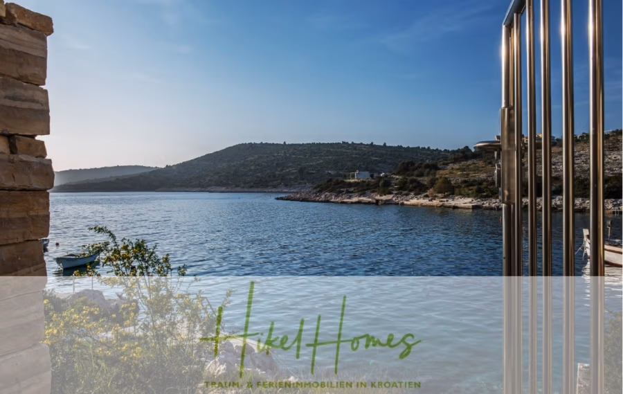 5 Sterne Luxus Villa Christina – 250m² WFL in 1. Reihe zum Meer/ 4 SZ / Neubau / Sofort bezugsfertig - Ein ruhiger Küstenblick mit ruhigem Wasser, das den blauen Himmel spiegelt. Felsige Hügel umgeben die Bucht, und ein kleines Boot liegt in Ufernähe vor Anker. Ein Metallgeländer und eine Steinmauer rahmen den Vordergrund ein. Textüberlagerung: „Hike Homes: Traum- & Ferienimmobilien in Kroatien.“. (41515)