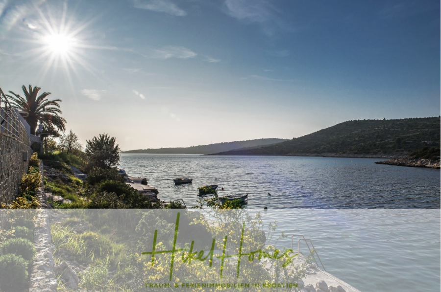 5 Sterne Luxus Villa Christina – 250m² WFL in 1. Reihe zum Meer/ 4 SZ / Neubau / Sofort bezugsfertig - Eine malerische Ansicht einer ruhigen Bucht mit kleinen Booten auf dem Wasser, einem Steinweg, üppigem Grün, einer Palme und Hügeln im Hintergrund unter einer hellen Sonne. Text Overlay am unteren Rand liest "Hakel Haus. (46286)