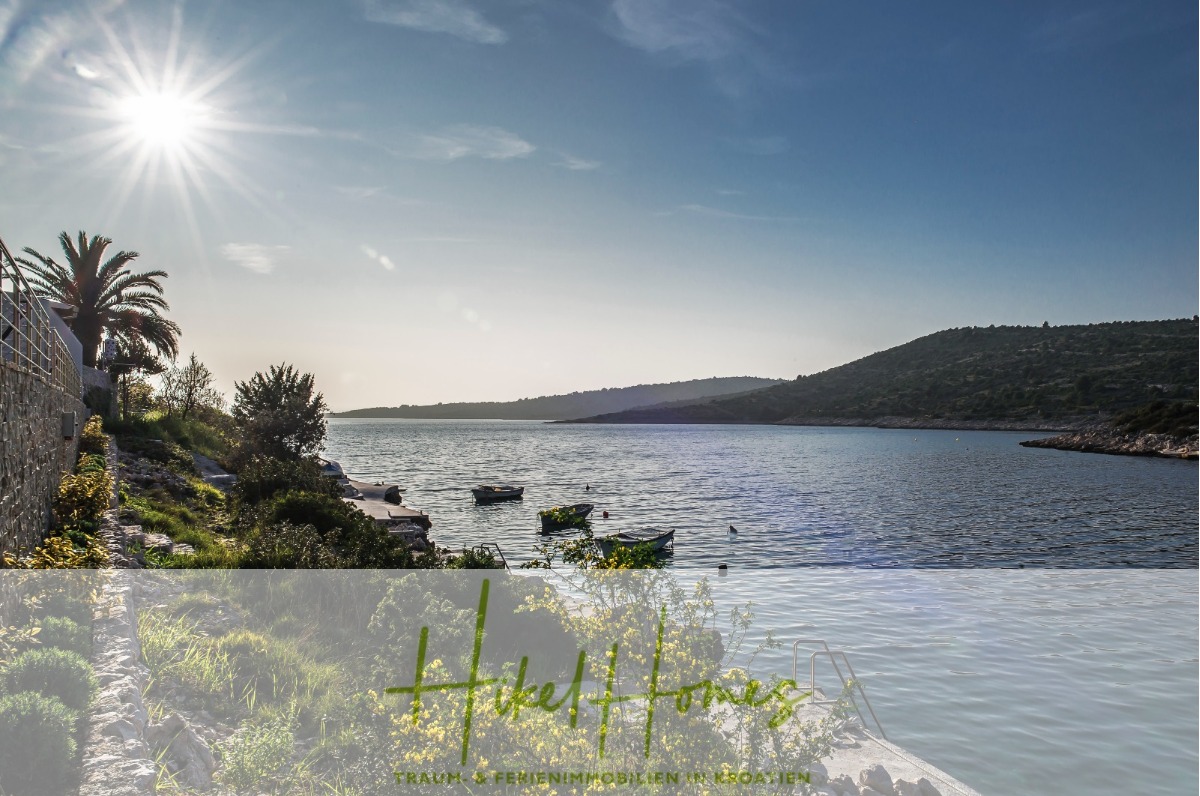 Eine malerische Ansicht einer ruhigen Bucht mit kleinen Booten auf dem Wasser, einem Steinweg, üppigem Grün, einer Palme und Hügeln im Hintergrund unter einer hellen Sonne. Text Overlay am unteren Rand liest "Hakel Haus. - 5 Sterne Luxus Villa Christina – 250m² WFL in 1. Reihe zum Meer/ 4 SZ / Neubau / Sofort bezugsfertig