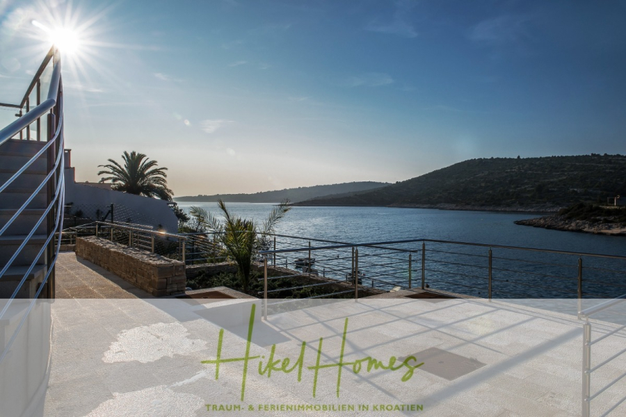 5 Sterne Luxus Villa Christina – 250m² WFL in 1. Reihe zum Meer/ 4 SZ / Neubau / Sofort bezugsfertig - Eine Terrasse am Wasser mit Metallgeländer bietet einen Blick auf eine ruhige Bucht und grüne Hügel unter einem klaren blauen Himmel bei Sonnenuntergang. Palmen und moderne Landschaftsgestaltung betonen die ruhige, malerische Umgebung. Der Text lautet: "Hikel Homes. Traum- & Ferienimmobilien in Kroatien. (46308)