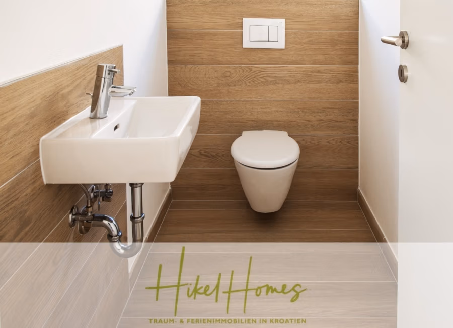 5 Sterne Luxus Villa Christina – 250m² WFL in 1. Reihe zum Meer/ 4 SZ / Neubau / Sofort bezugsfertig - Minimalistisches Badezimmer mit wandhängender Toilette und rechteckigem Waschbecken. Helle Holzvertäfelungen säumen Wände und Boden. Rechts ist eine teilweise geöffnete Tür zu sehen. Unten steht „Hikel Homes“ mit kleinem Text in einer Fremdsprache. (41518)