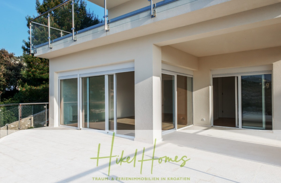 5 Sterne Luxus Villa Christina – 250m² WFL in 1. Reihe zum Meer/ 4 SZ / Neubau / Sofort bezugsfertig - Ein modernes Haus mit großen Glasschiebetüren, die auf eine große, geflieste Terrasse führen. Das Obergeschoss hat ein gläsernes Balkongeländer, und rund um das Haus sind Bäume und Grünflächen zu sehen. (46294)