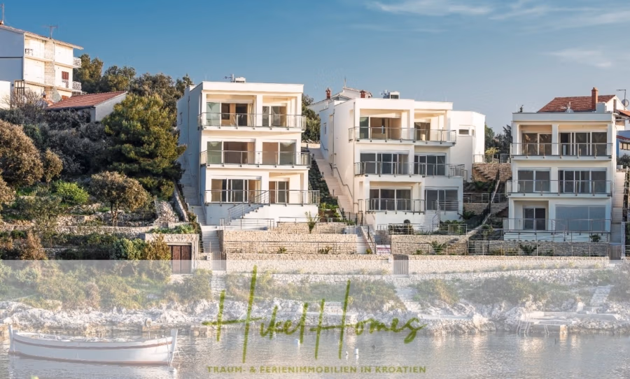 5 Sterne Luxus Villa Christina – 250m² WFL in 1. Reihe zum Meer/ 4 SZ / Neubau / Sofort bezugsfertig - Drei moderne weiße Villen an einem Hang blicken auf ein Gewässer. Kleine Boote liegen am felsigen Ufer. Umgeben von Bäumen und anderen Häusern verfügen die Villen über Balkone und große Fenster. Der Schriftzug „Dream & Ferienimmobilien in Kroatien“ ist sichtbar. (41498)