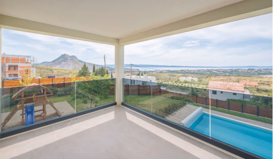 Villa am Stadtrand von Split großzügige Aufteilung, hohe Räume und beheiztem Pool - Geräumiger Balkon mit Glasgeländer mit malerischer Aussicht auf Berge, Felder und ein Gewässer in der Ferne. Darunter befinden sich ein Swimmingpool und ein Kinderspielplatz mit Schaukel. (41933)