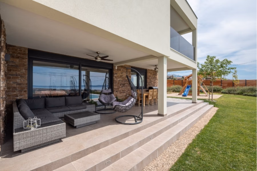 Villa am Stadtrand von Split großzügige Aufteilung, hohe Räume und beheiztem Pool - Moderne Terrasse mit grauem Sofa, Rattanmöbeln und Hängesesseln auf einer gefliesten Veranda. Angrenzender Barbereich mit Hockern. Im Hintergrund: grüner Rasen, Holzspielgeräte und teilweise bewölkter Himmel. (41929)